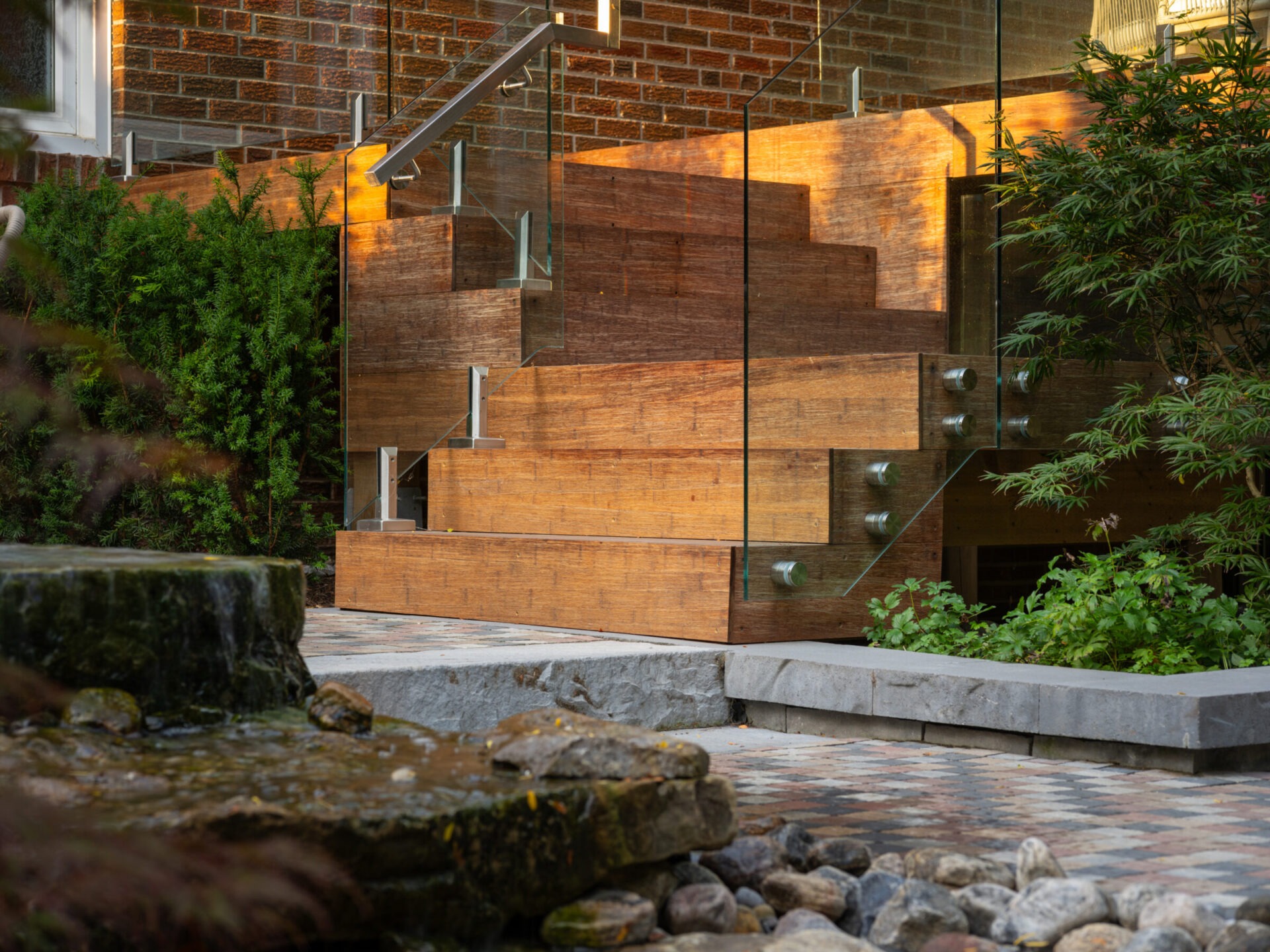 Modern outdoor staircase with wooden steps and glass railing, surrounded by greenery and a small stone-lined pond in a tranquil garden setting.