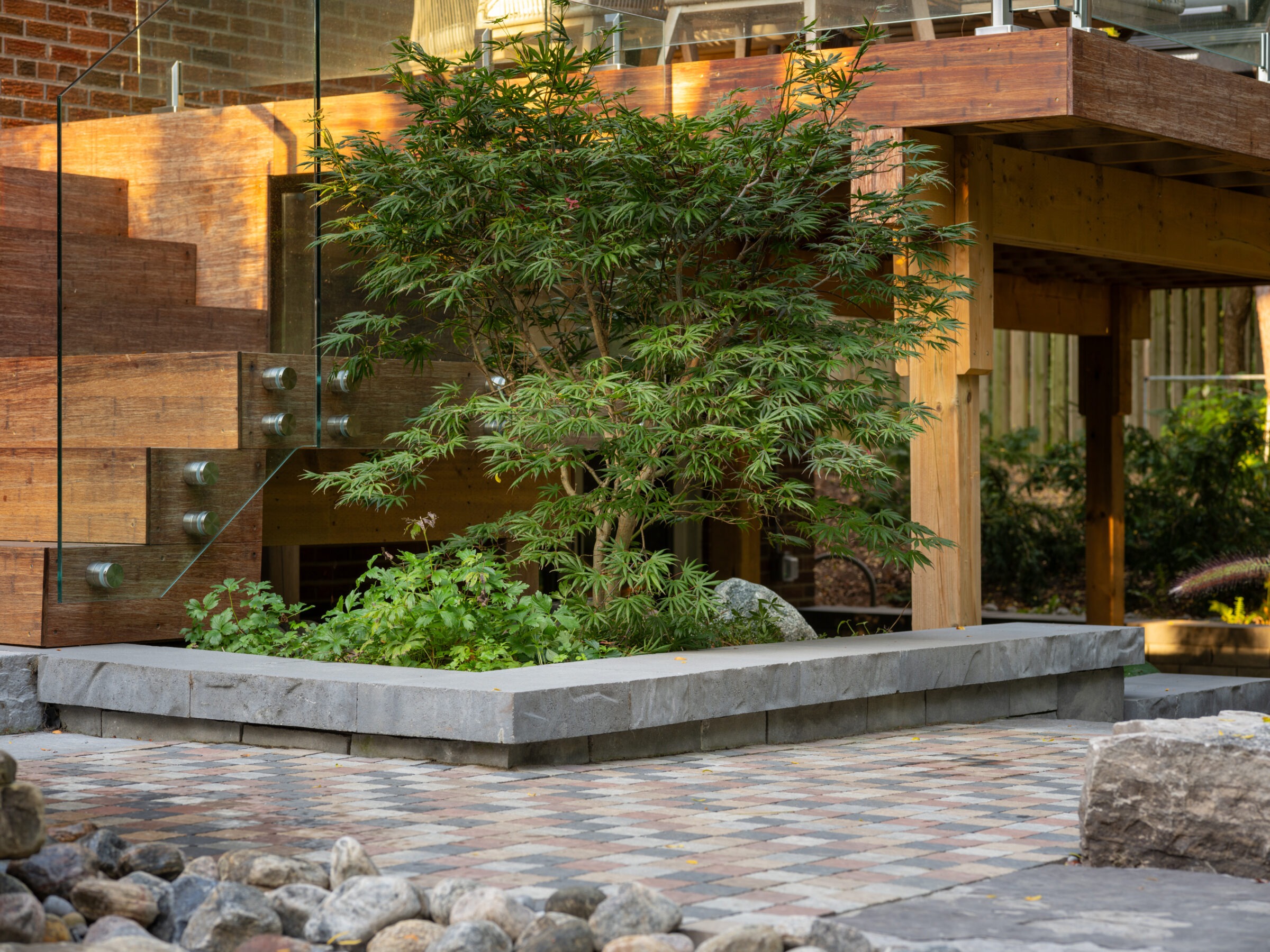 Modern outdoor space with interlocking stone patio, wooden deck, glass railing, and lush greenery featuring small tree and various plants. Tranquil garden setting.