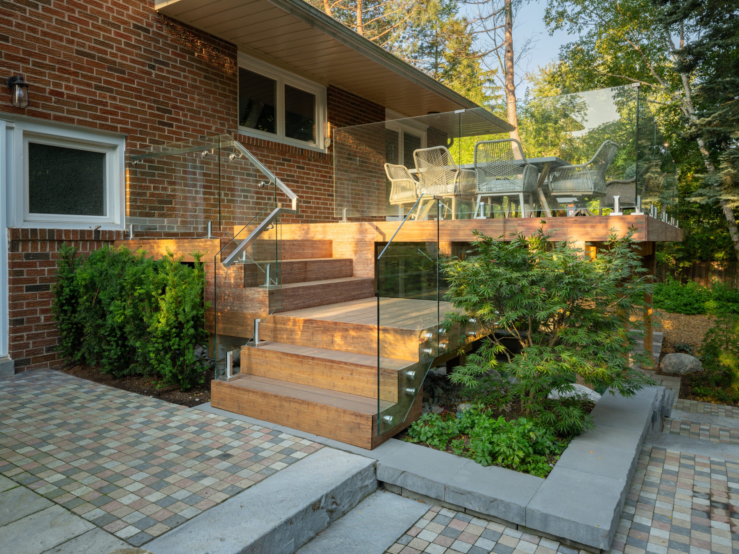 Brick house with glass-railed wooden deck, outdoor seating, and lush greenery. Stone paving and steps lead to vibrant garden landscaping.