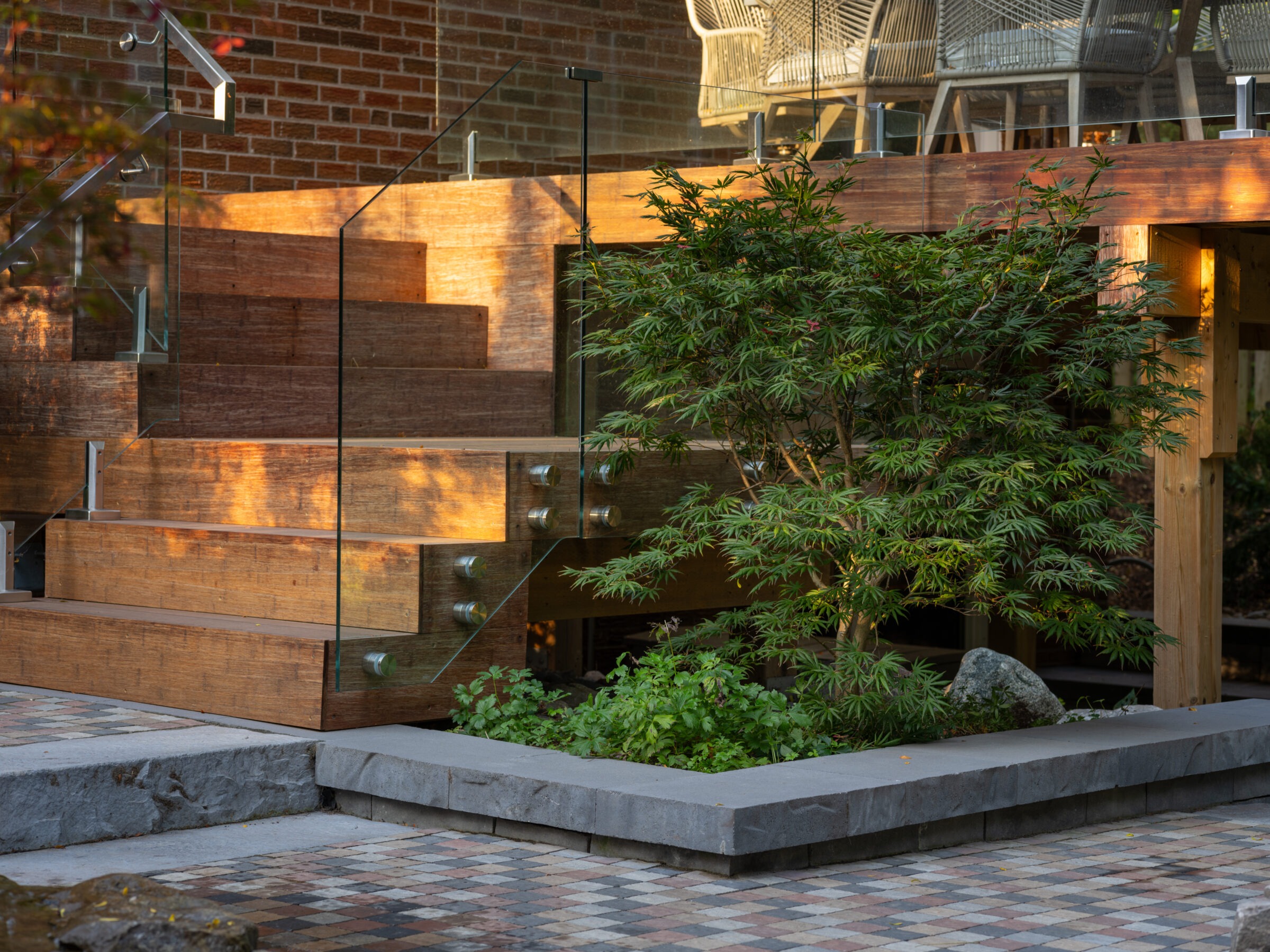 Modern outdoor patio with wooden stairs, glass railing, and lush green foliage. Sunlight highlights the brick wall and checkered stone floor.