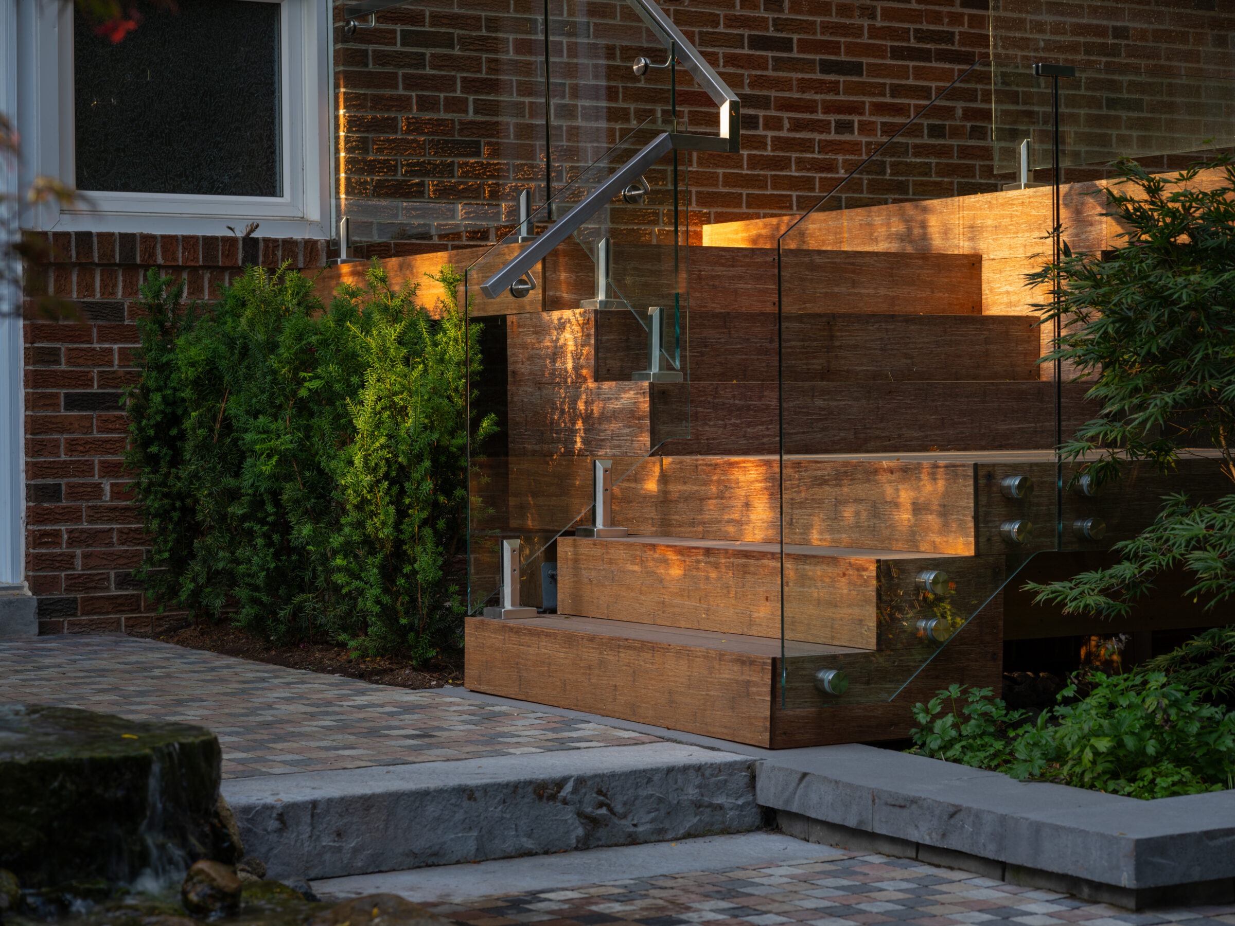 Modern brown brick building entrance with wooden steps, glass railings, and greenery. Sunlight casts warm glow, enhancing the architectural design.
