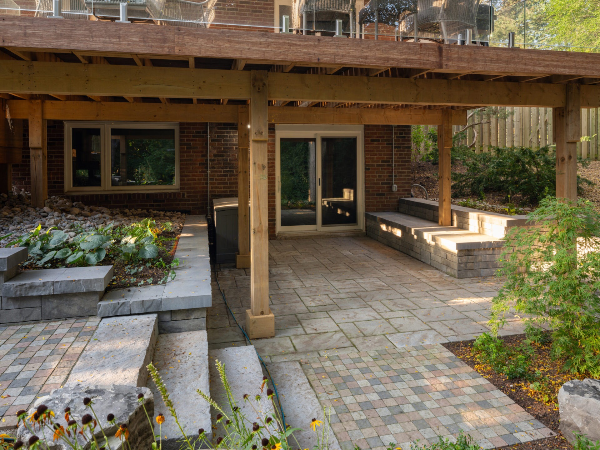 A wooden pergola over a stone patio with lush greenery and sliding glass doors of a brick house in a serene backyard setting.