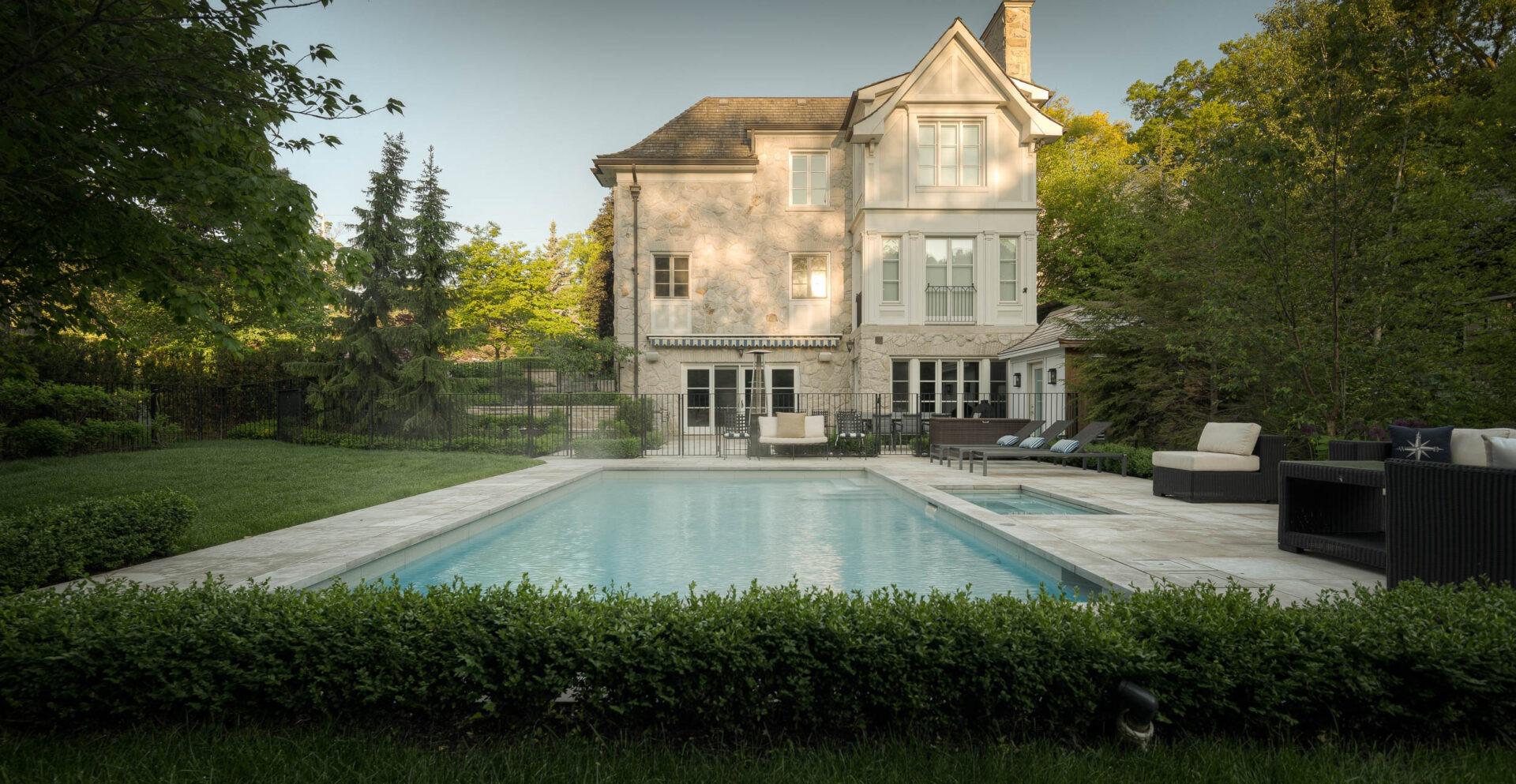 Formal backyard with a rectangular swimming pool, stone pool deck, and outdoor seating set in front of a large stone house surrounded by mature trees and lawn.