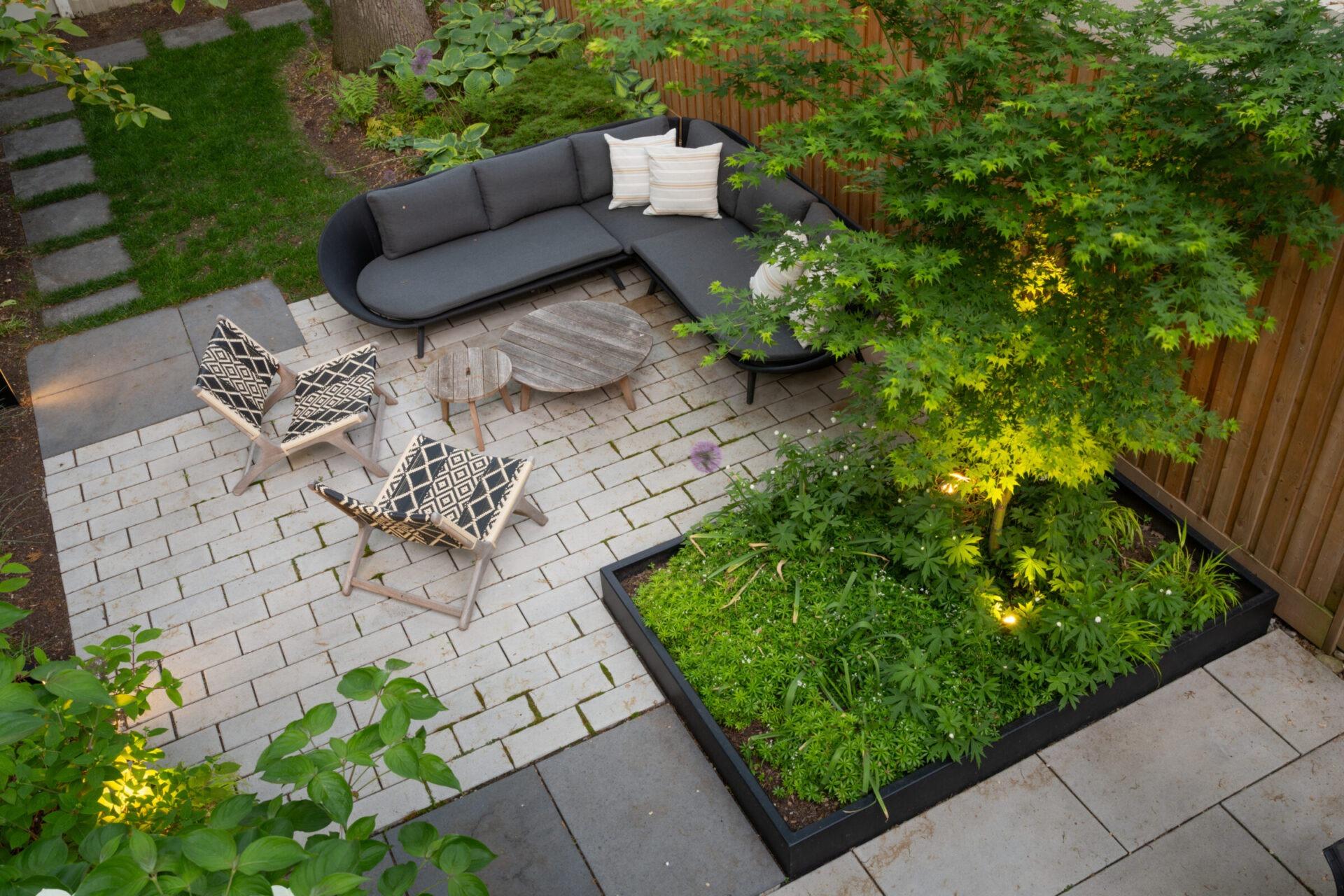 Overhead view of a modern patio with a curved outdoor sofa, patterned lounge chairs, wood coffee tables, and a raised planter with a Japanese maple and soft landscape lighting.