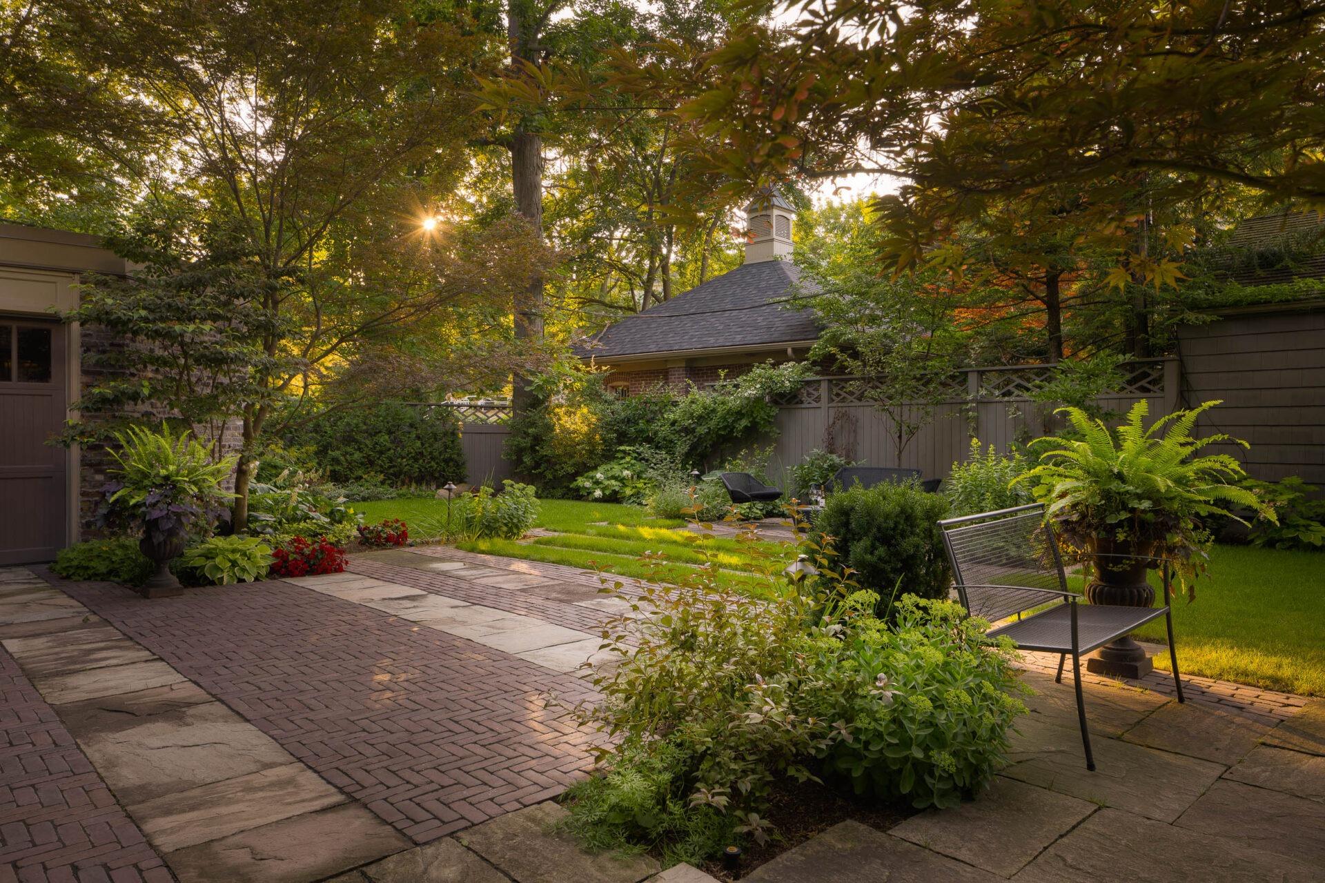 Shaded garden courtyard with stone and brick pathways, lush planting beds, outdoor seating, and mature trees filtering warm evening light.