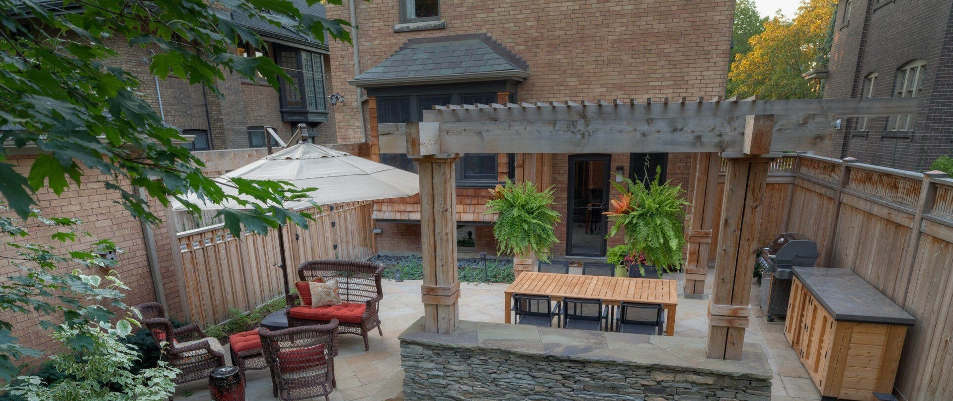 Cozy backyard patio with wooden pergola, wicker chairs, table, and grill. Lush green plants and brick walls create a serene outdoor space.